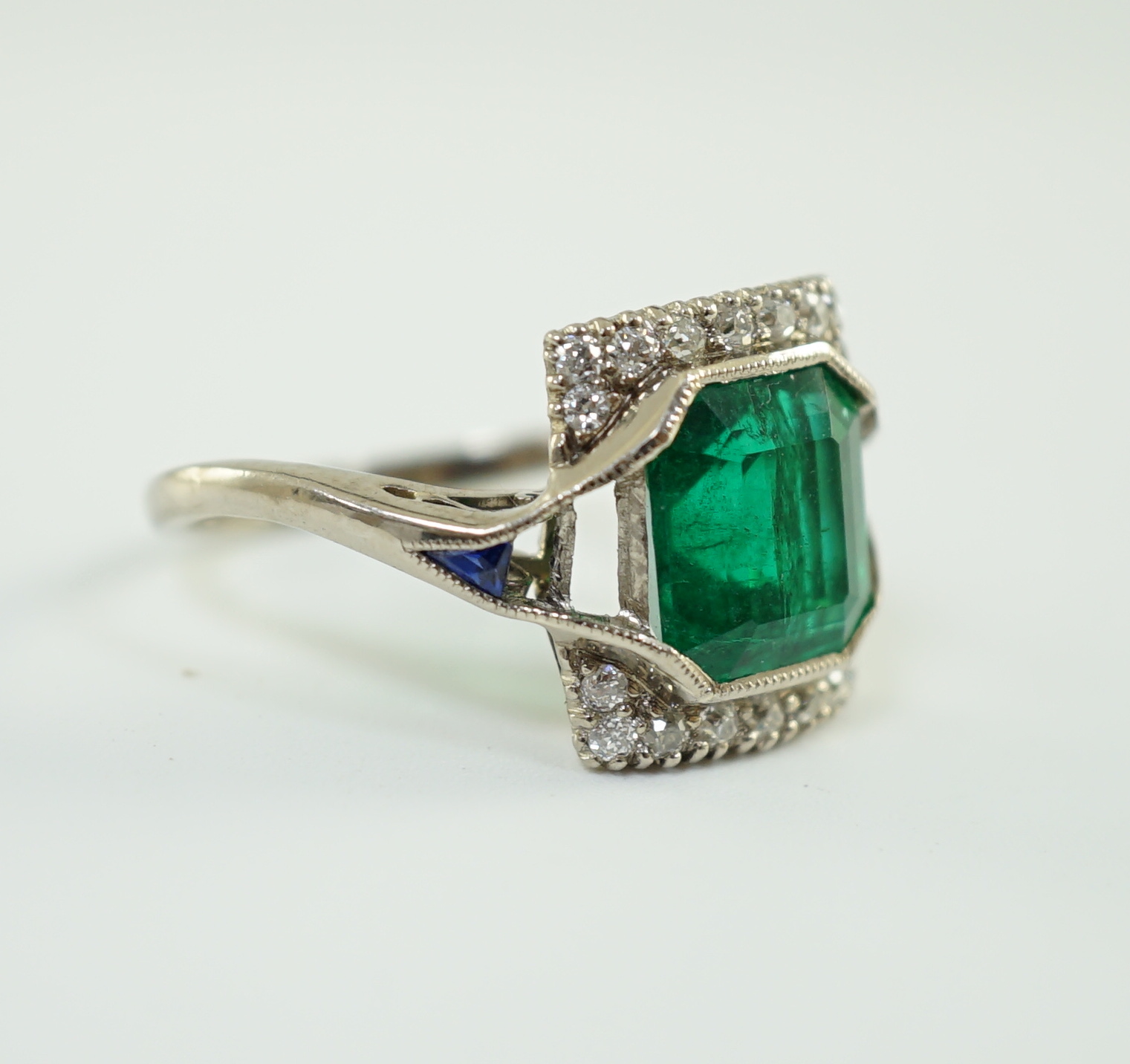 An Art Deco 18k white gold, emerald and diamond set square cluster dress ring, with shaped cut sapphire set shoulders, (a.f.)